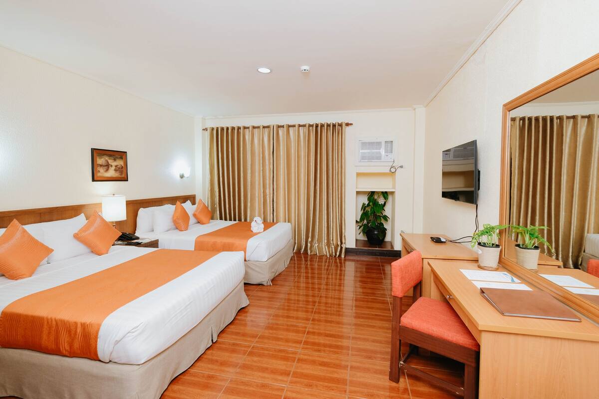 A hotel room with two beds, a desk, a chair, a mirror, a TV, and a potted plant.
