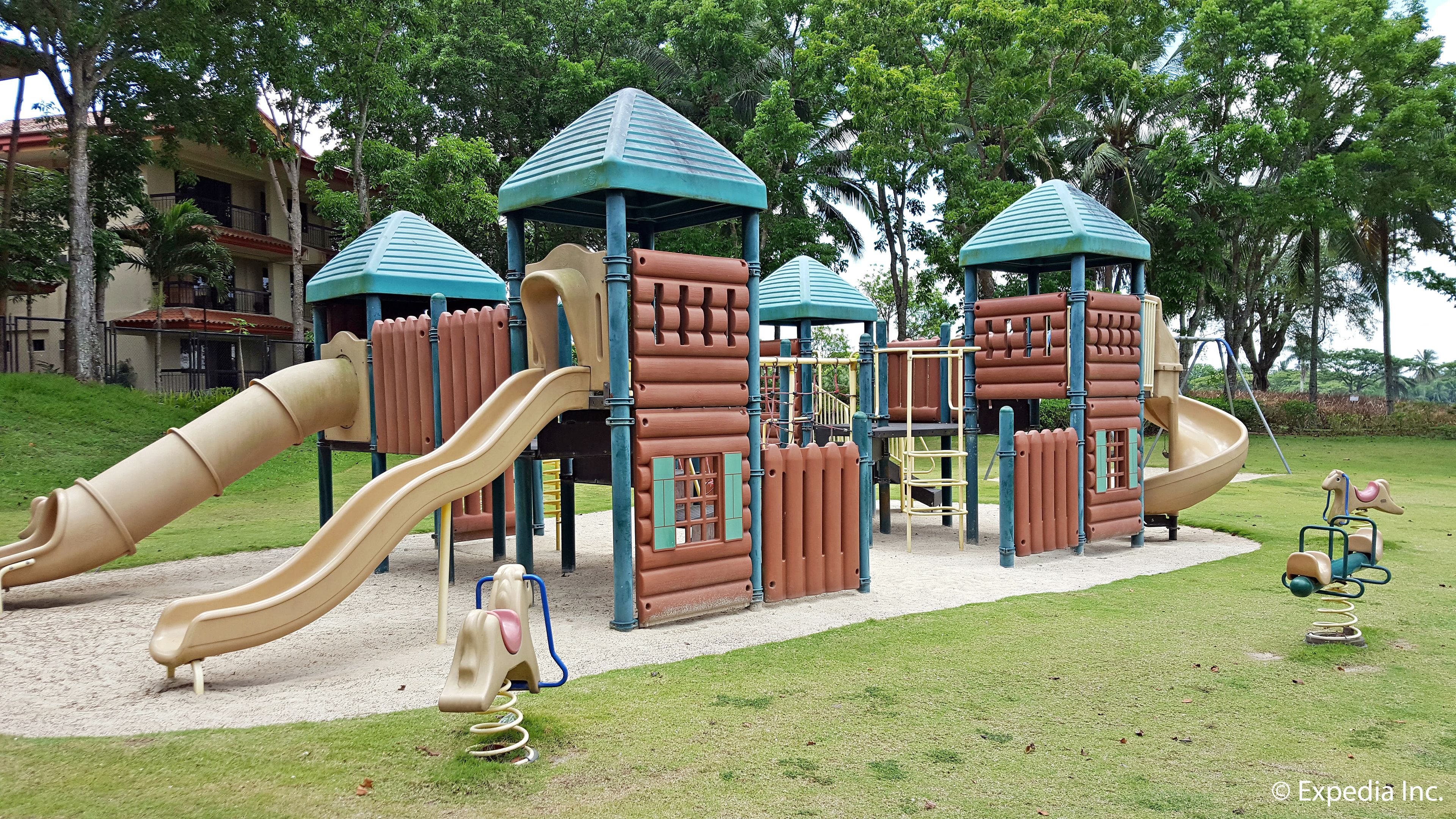 A playground with slides and climbing structures, surrounded by grass and trees.