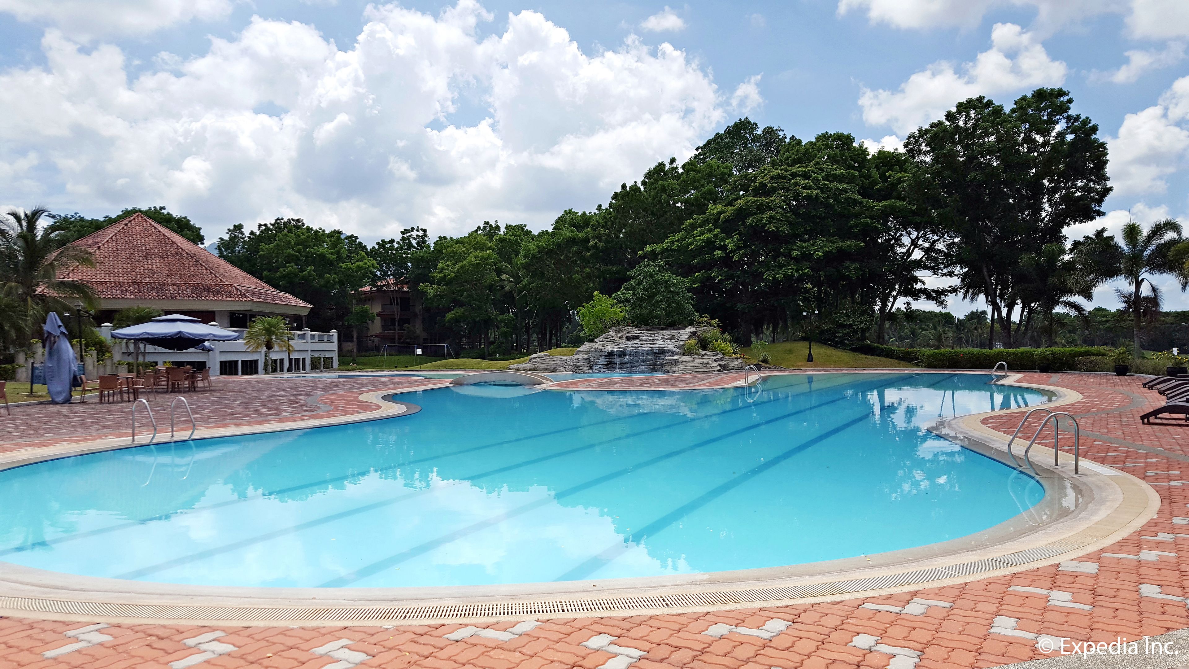 Una piscina con cascada, rodeada por un patio de ladrillo y zonas de estar a la sombra.