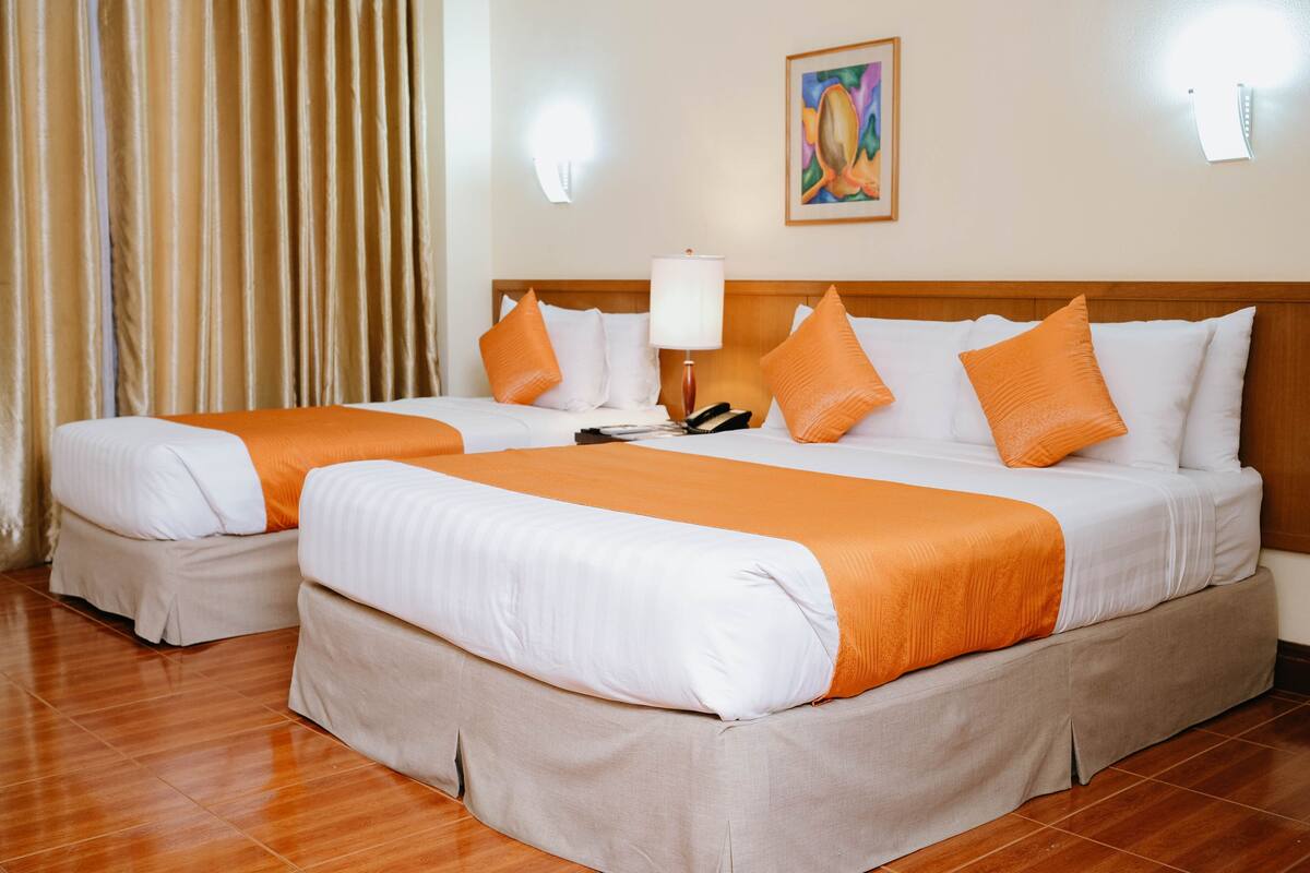 A hotel room with two beds, beige and orange bedding, a wooden headboard, a painting on the wall, and a lamp.