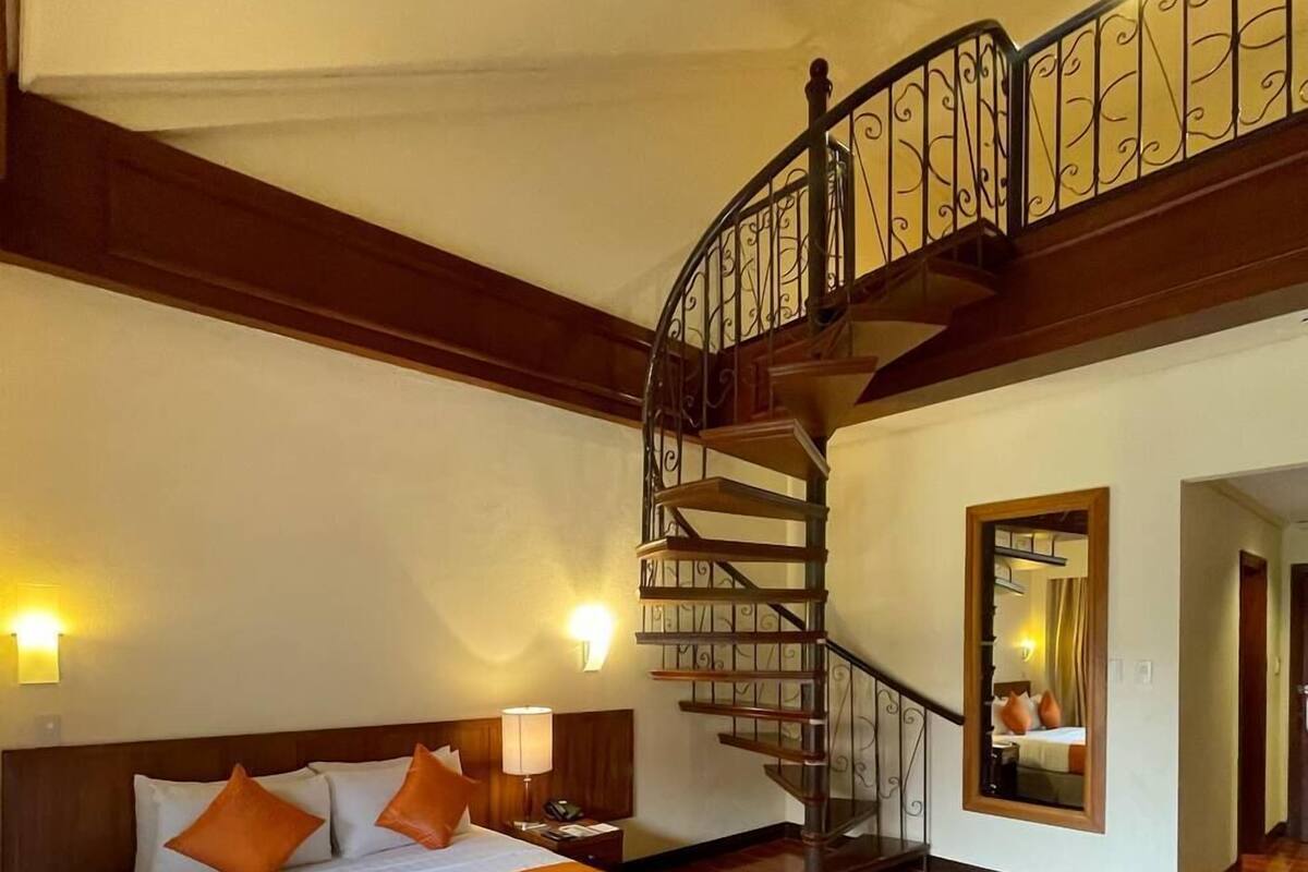 A spacious bedroom with a large bed, a wooden staircase, and a mirror on the wall.
