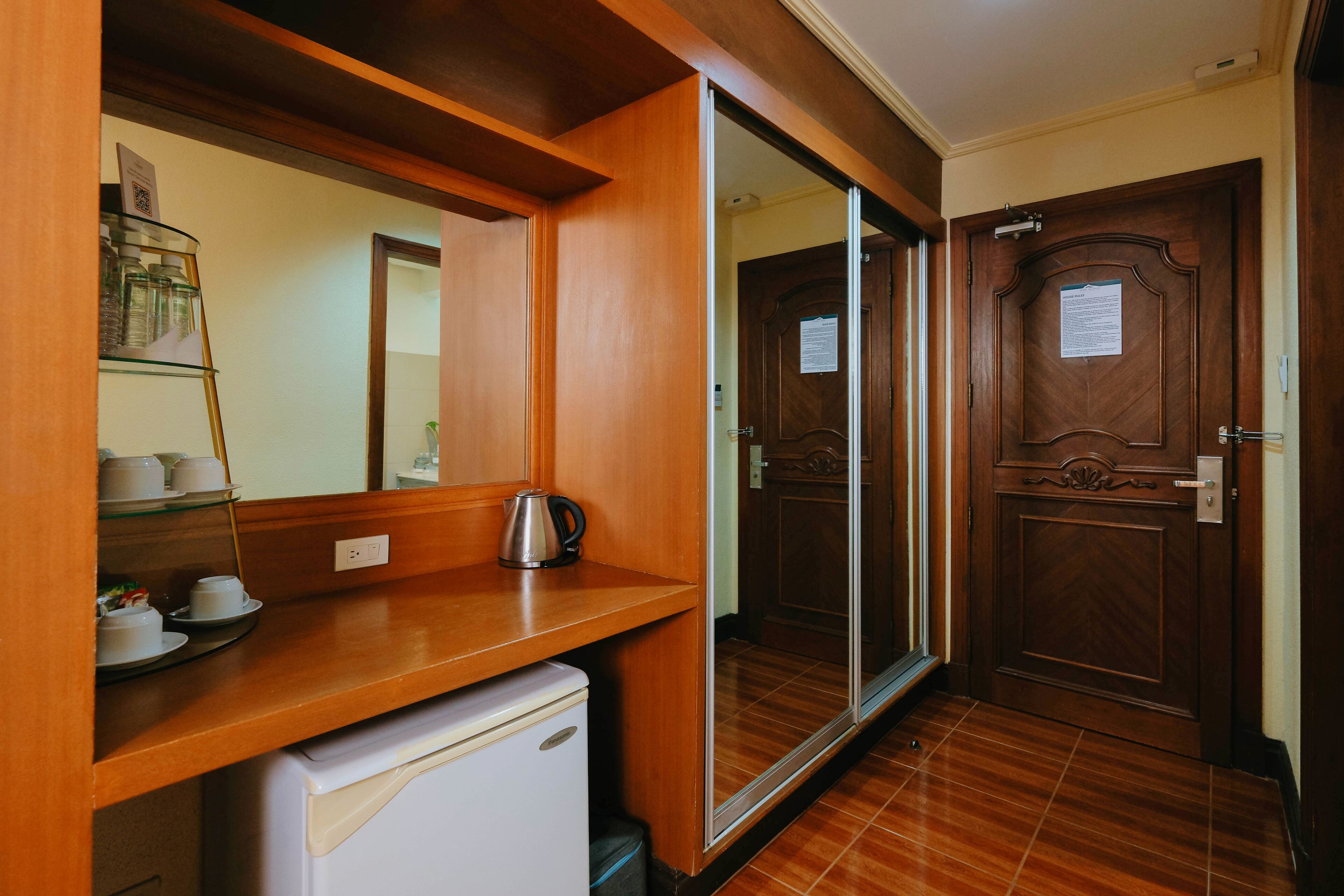 Una kitchenette compacta con refrigerador, cafetera y repisas de vidrio con tazas y hervidor.