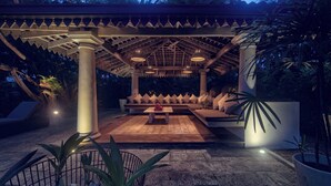 Un espacio lounge al aire libre con un conjunto de asientos cubierto, una mesa central y plantas en macetas.