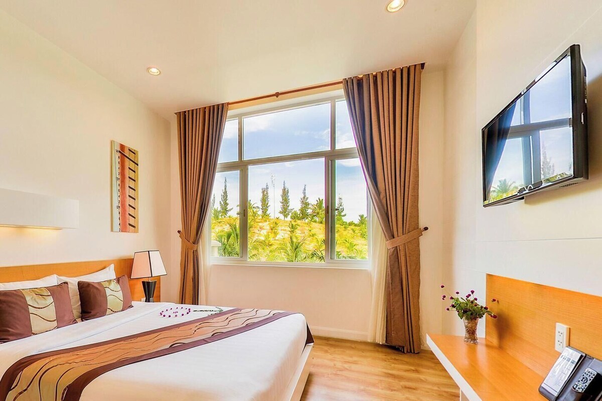 A hotel room with a bed, a desk, a TV, a window with curtains, and a view of greenery outside.