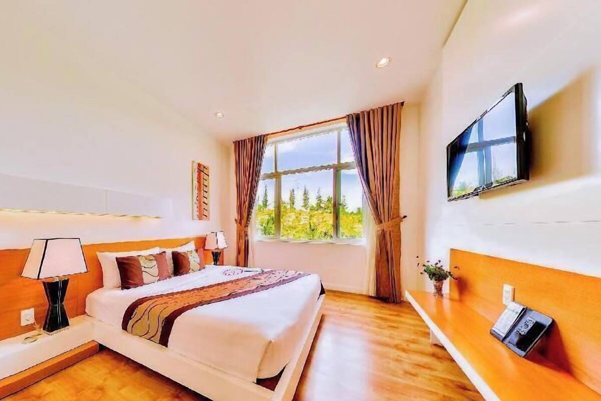Habitación de hotel con cama, mesitas de noche, televisión, ventana con cortinas y vistas a árboles.
