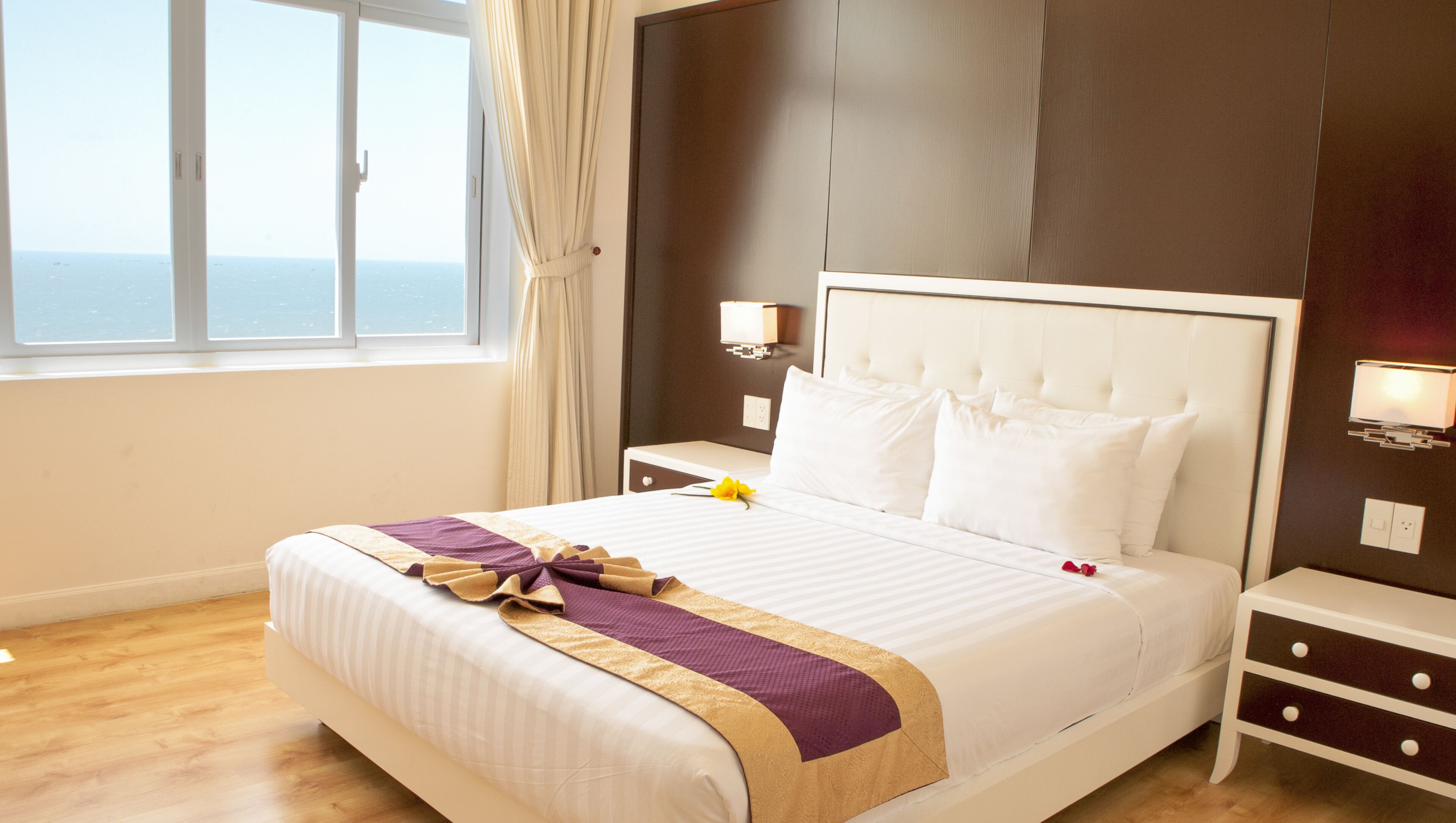 Habitación de hotel con una cama grande, una mesita de noche, una lámpara de pared y vistas al mar a través de una ventana.