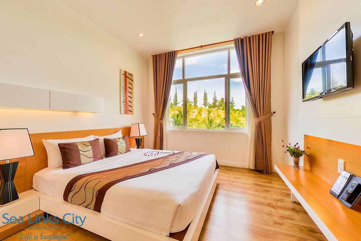 Habitación de hotel con una cama grande, un televisor en la pared, una ventana con cortinas y suelo de madera.