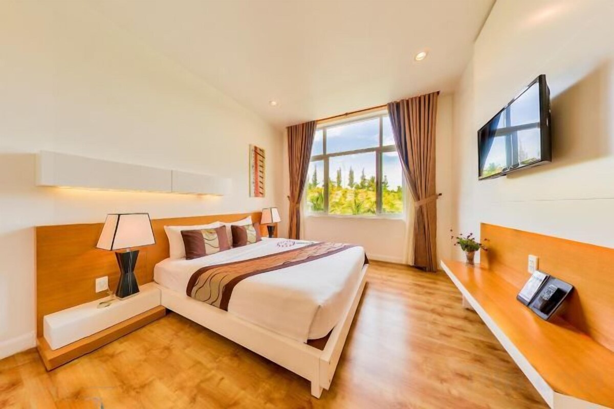 A hotel room with a bed, bedside table, lamp, TV, window, and a view of greenery outside.