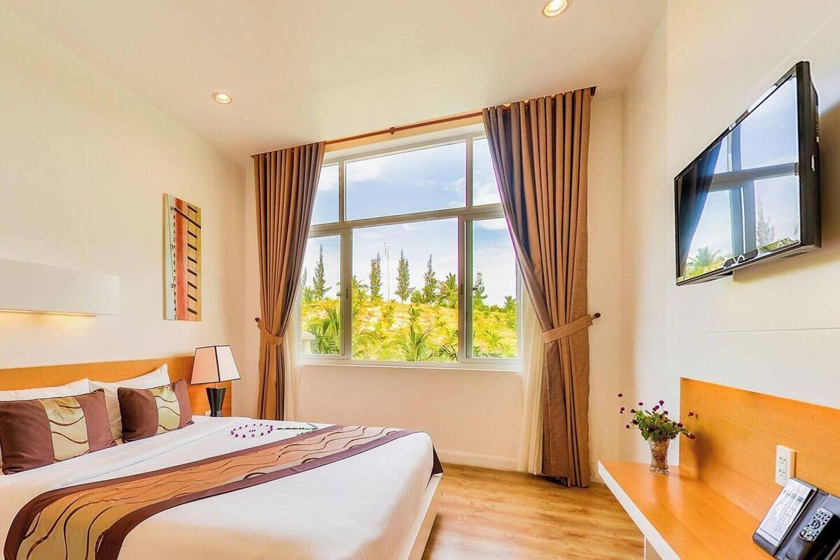 Habitación de hotel con cama, escritorio, televisión, ventana con cortinas y vista a zonas verdes.