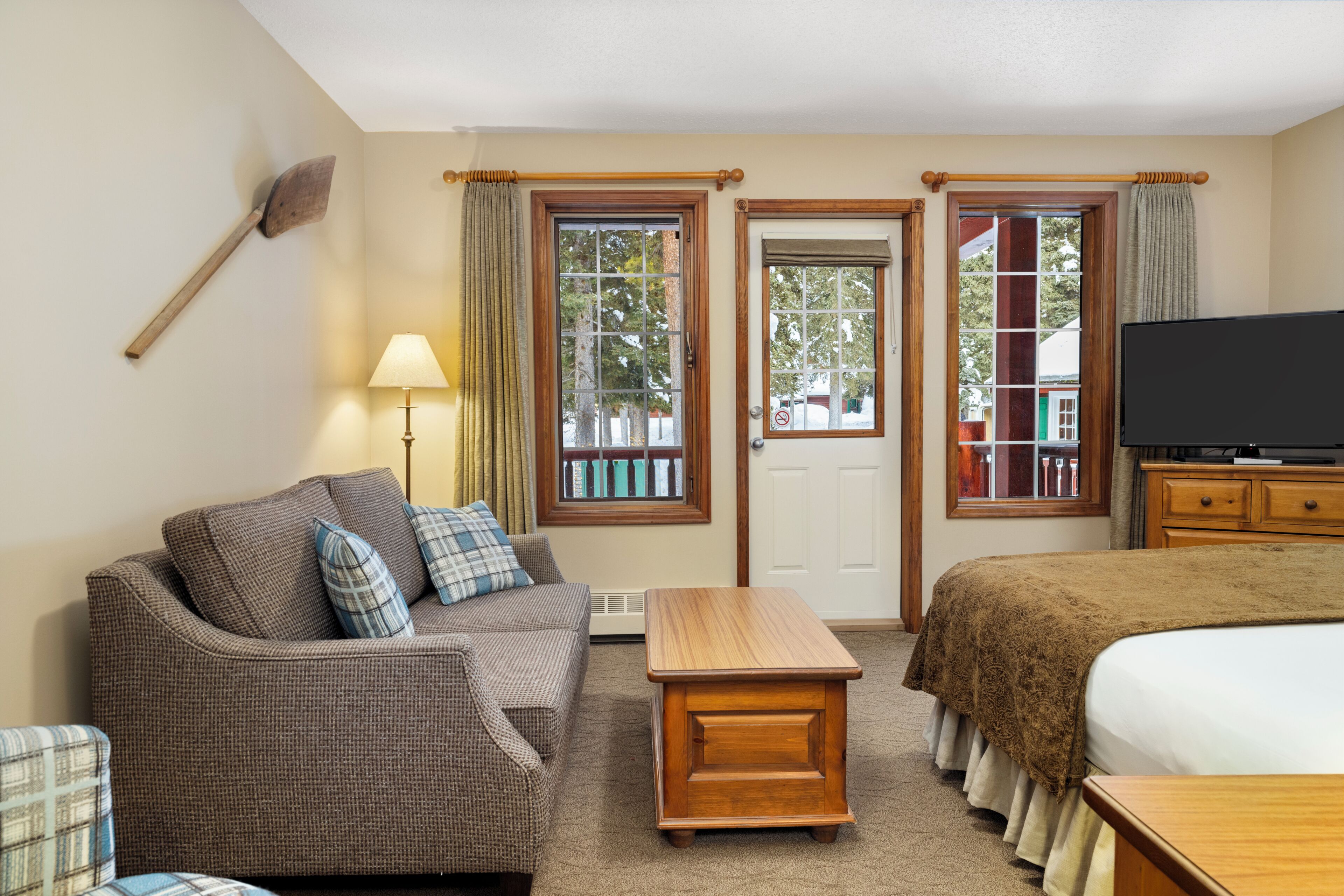 A hotel room with a sofa, a wooden table, a bed, a television, and two windows with curtains.