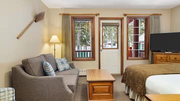 A hotel room with a sofa, a wooden table, a bed, a television, and two windows with curtains.