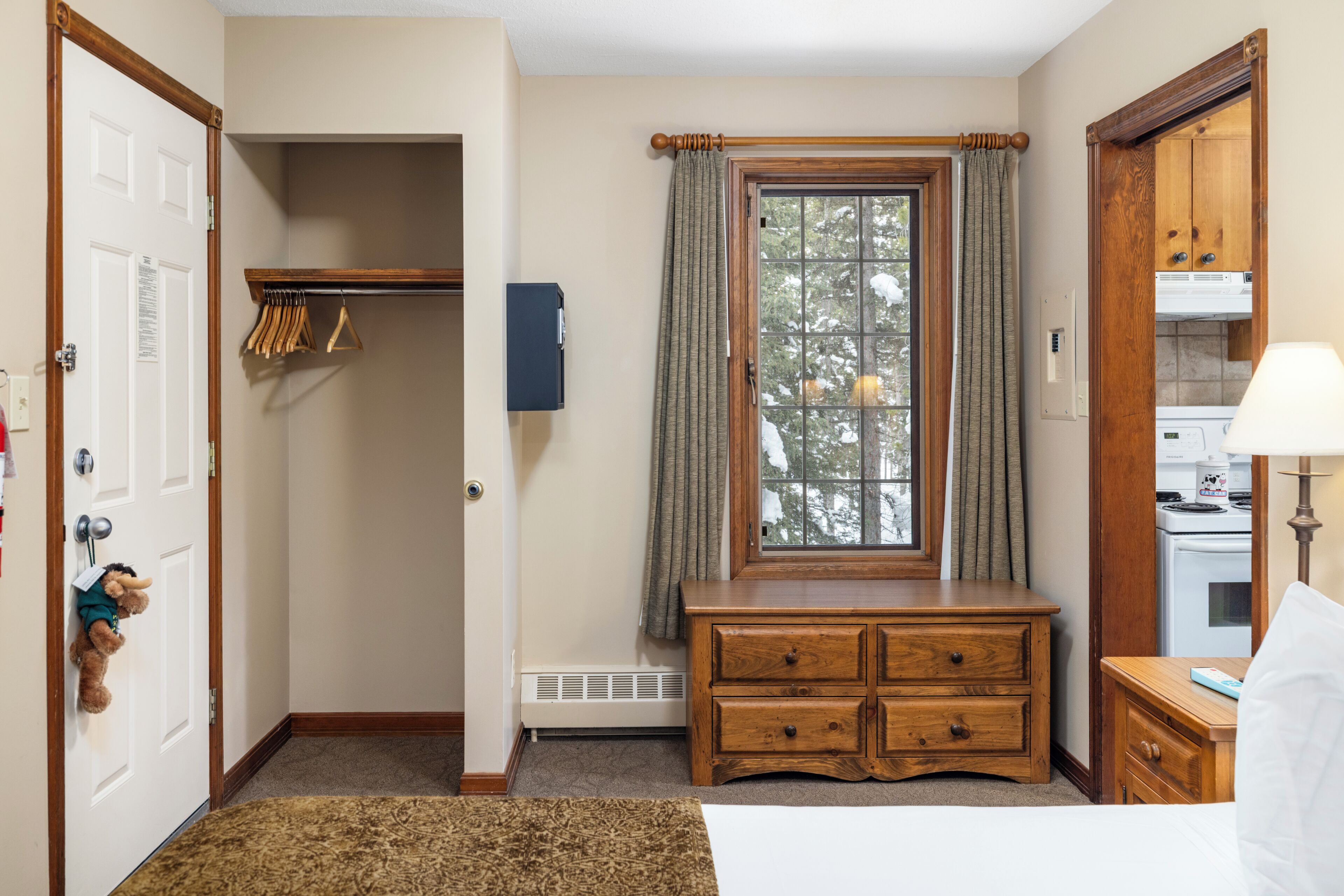 Um quarto com um banco de madeira, um radiador, uma janela com cortinas, um porta-roupas e uma porta com um brinquedo macio.