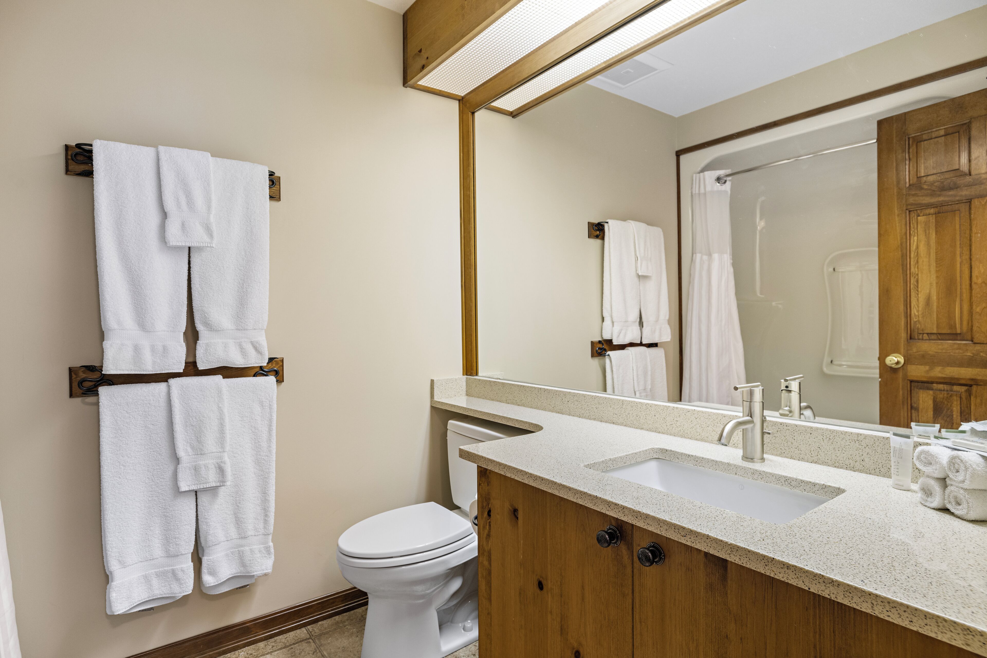 Un baño con inodoro, lavamanos y espejo. Hay toallas colgadas en ganchos.