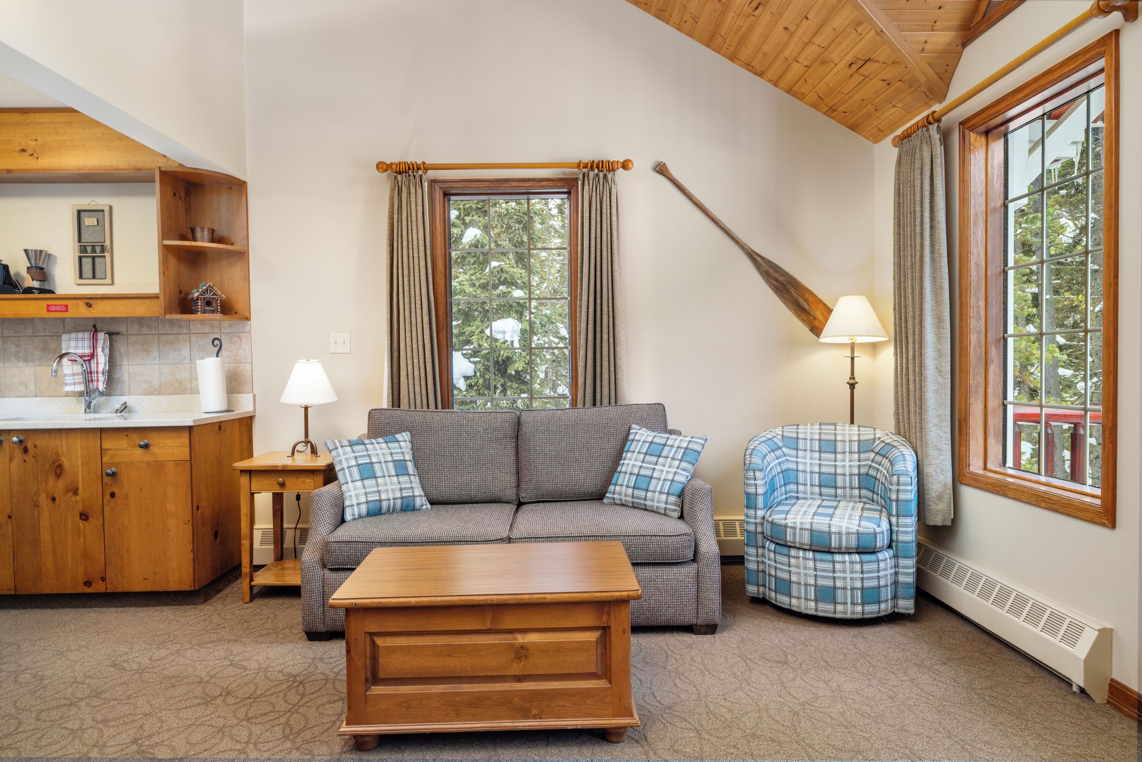 A cozy living room with a sofa, a chair, a coffee table, and a lamp. There is a window with curtains and a view of trees outside.
