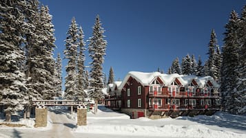 Uma pousada com entrada de pedra, rodeada por árvores cobertas de neve, e um céu azul límpido.