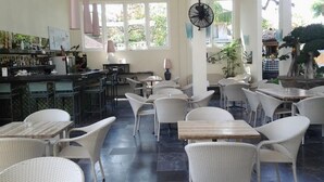 Un restaurante con mobiliario blanco de mimbre, una zona de bar y ventanas amplias que dejan entrar la luz natural.