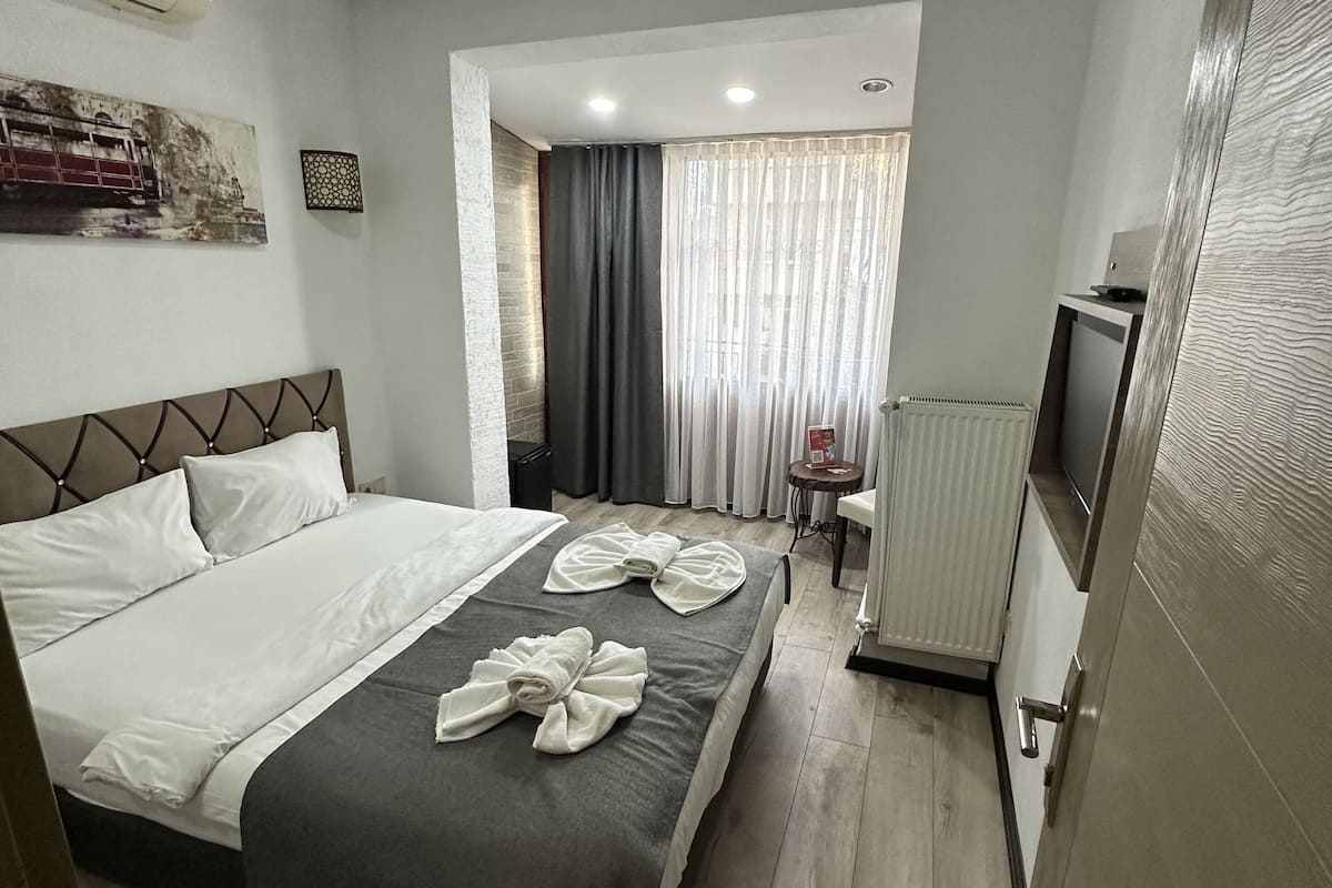 A hotel room with a bed, a TV, a radiator, and a window with curtains.