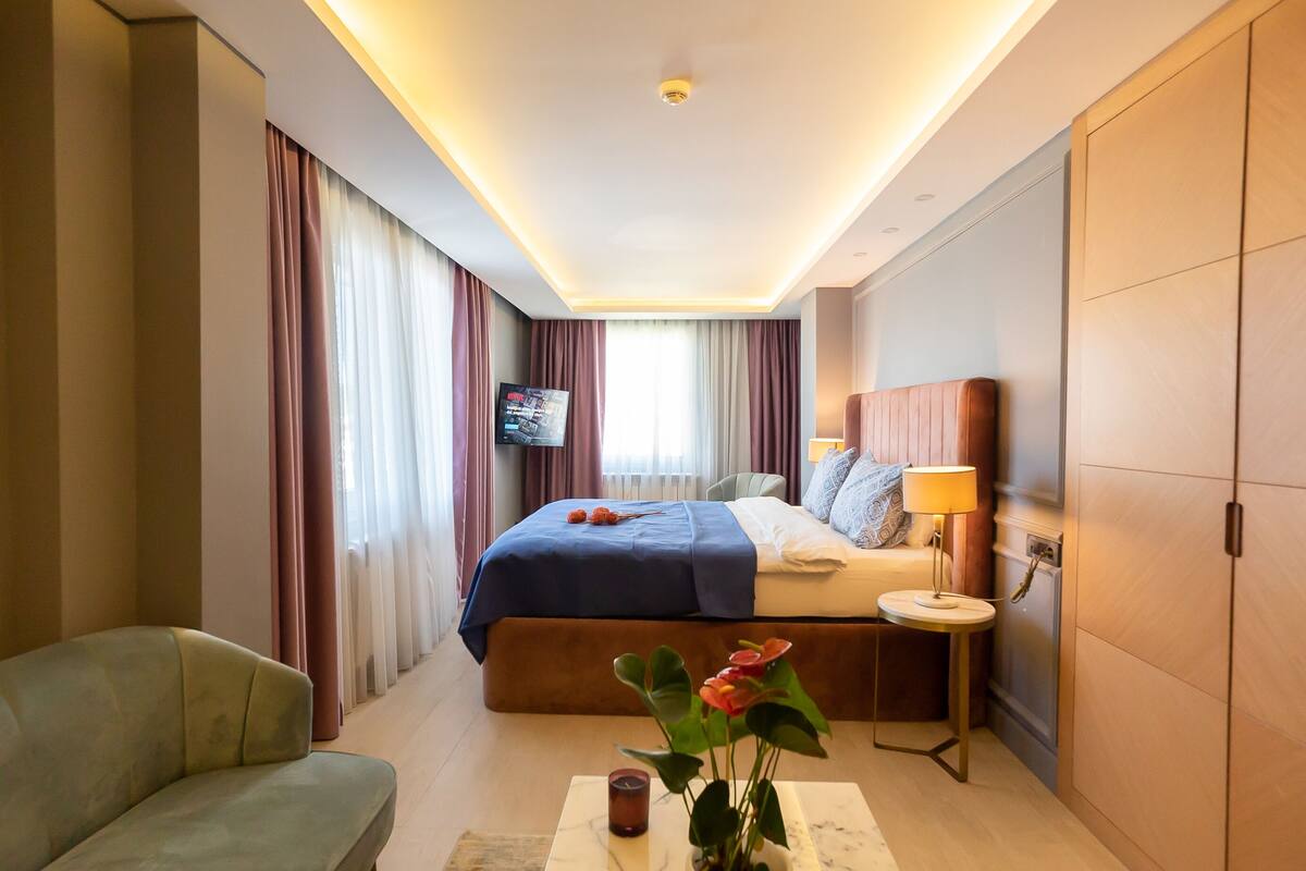 Habitación de hotel con cama, televisión, mesita de noche con lámpara, sofá y un florero con flores.