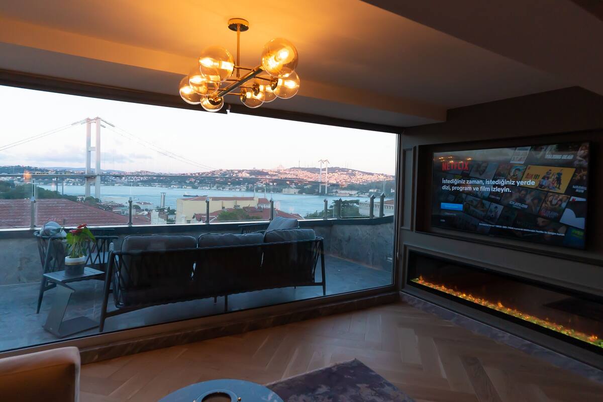 Una sala de estar moderna con un televisor grande, una chimenea y un balcón con vistas a un puente y al paisaje urbano.