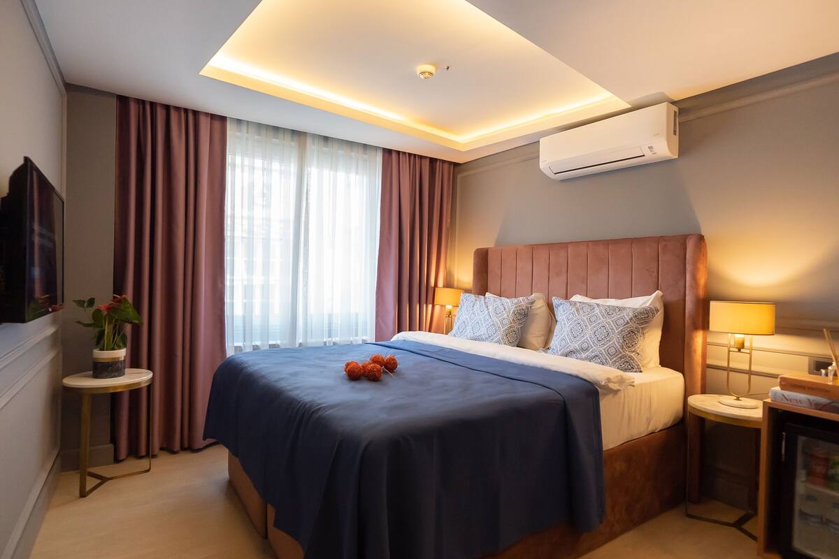 Una habitación de hotel moderna con una cama grande, un televisor de pantalla plana, una mesita de noche con lámpara y un equipo de aire acondicionado.