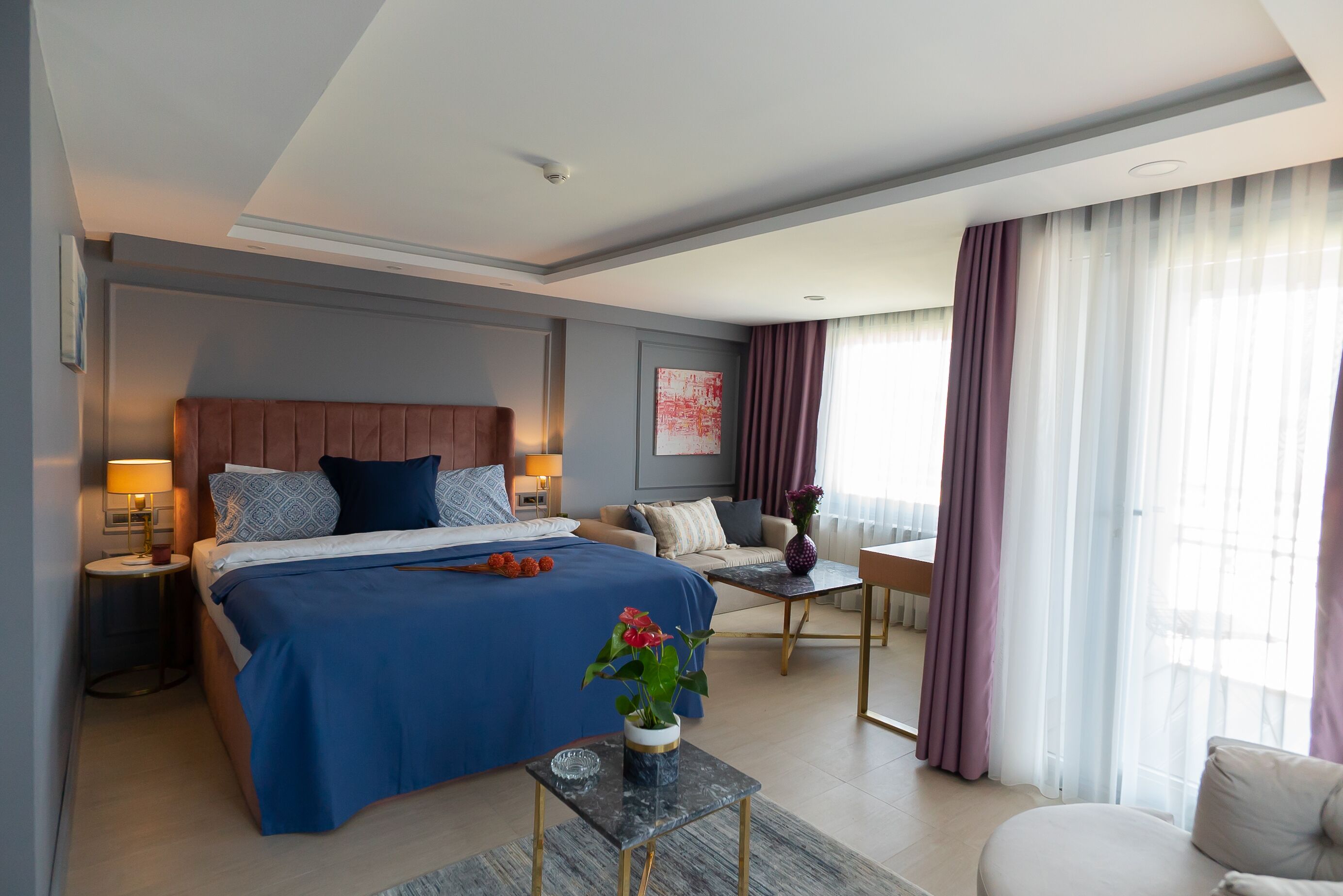 A modern hotel room with a large bed, a sitting area with a sofa and armchairs, a small table, and a large window with curtains.