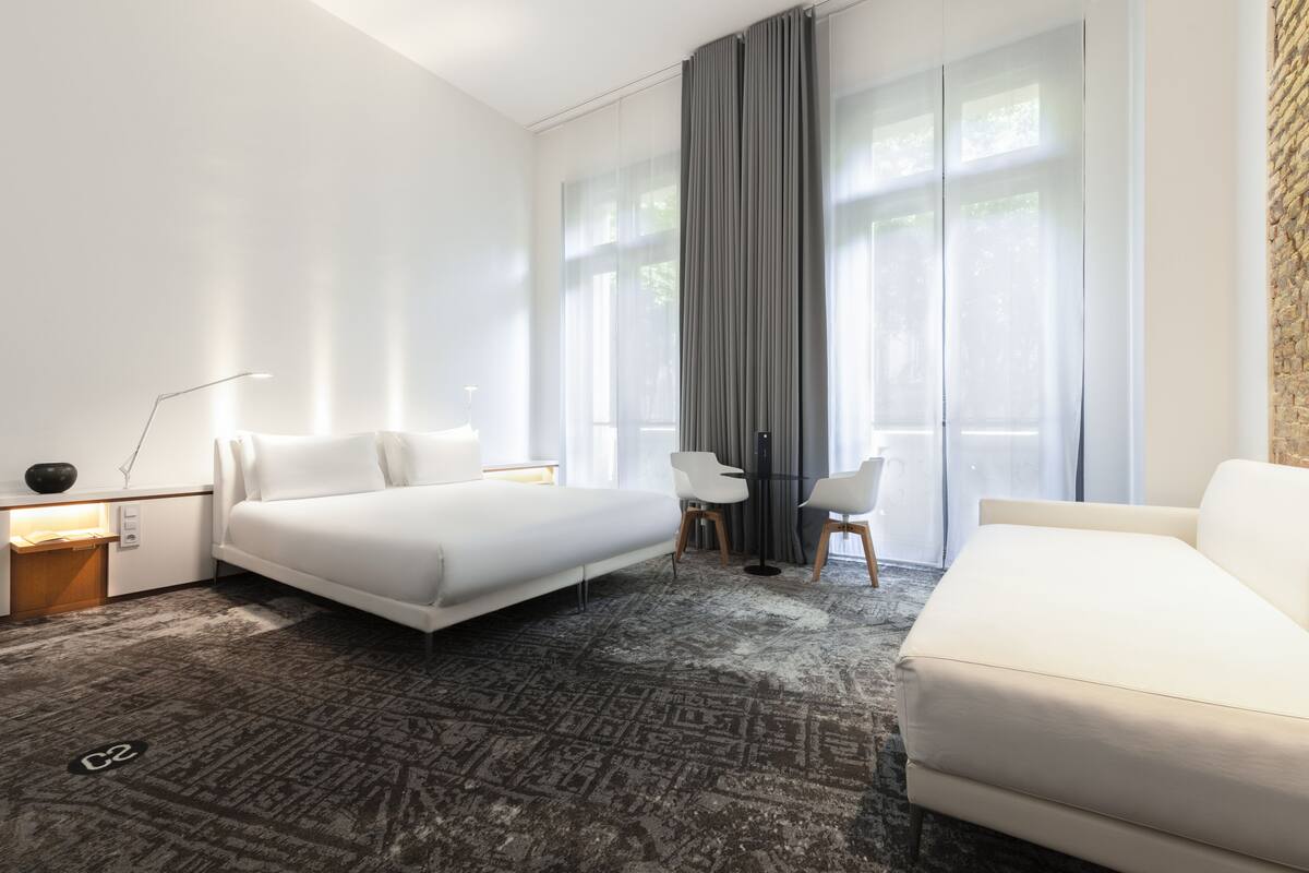 Une chambre d’hôtel moderne avec un grand lit, un canapé, un bureau avec deux chaises, une lampe et une grande fenêtre avec des rideaux.