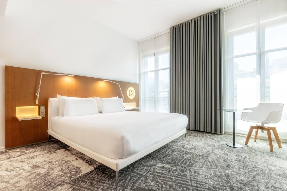 Une chambre d’hôtel moderne dotée d’un grand lit, de lampes de chevet, d’un bureau et offrant une vue sur la ville grâce à de grandes fenêtres.