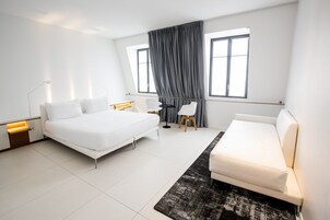 Habitación de hotel moderna con una cama grande, un sofá, un escritorio y una silla. Dos ventanas con cortinas, una lámpara de pie y una alfombra.