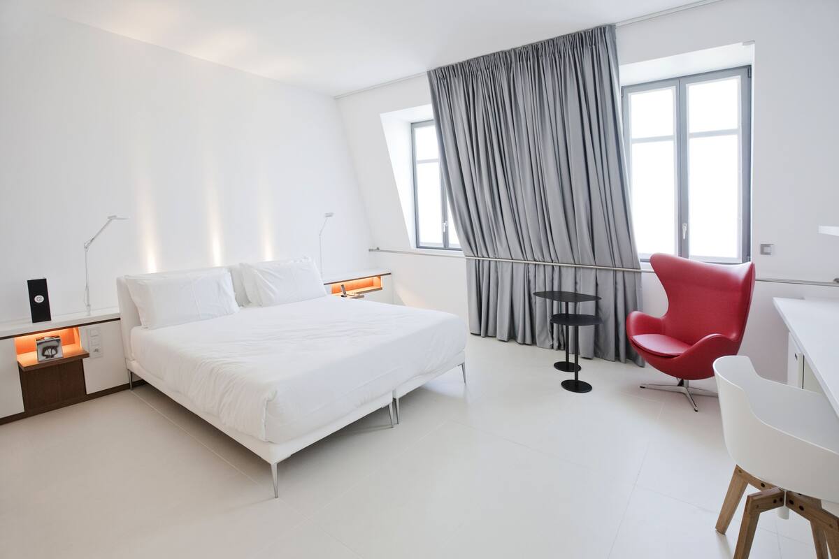 Une chambre d’hôtel moderne avec un grand lit, un fauteuil rouge, un bureau et une fenêtre avec des rideaux.