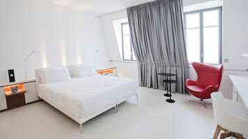Habitación de hotel moderna con una cama grande, una silla roja, un escritorio y un ventanal con cortinas.