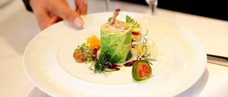 A white plate with a green vegetable-based dish, garnished with herbs and a small piece of meat, accompanied by a fork and a wine glass.