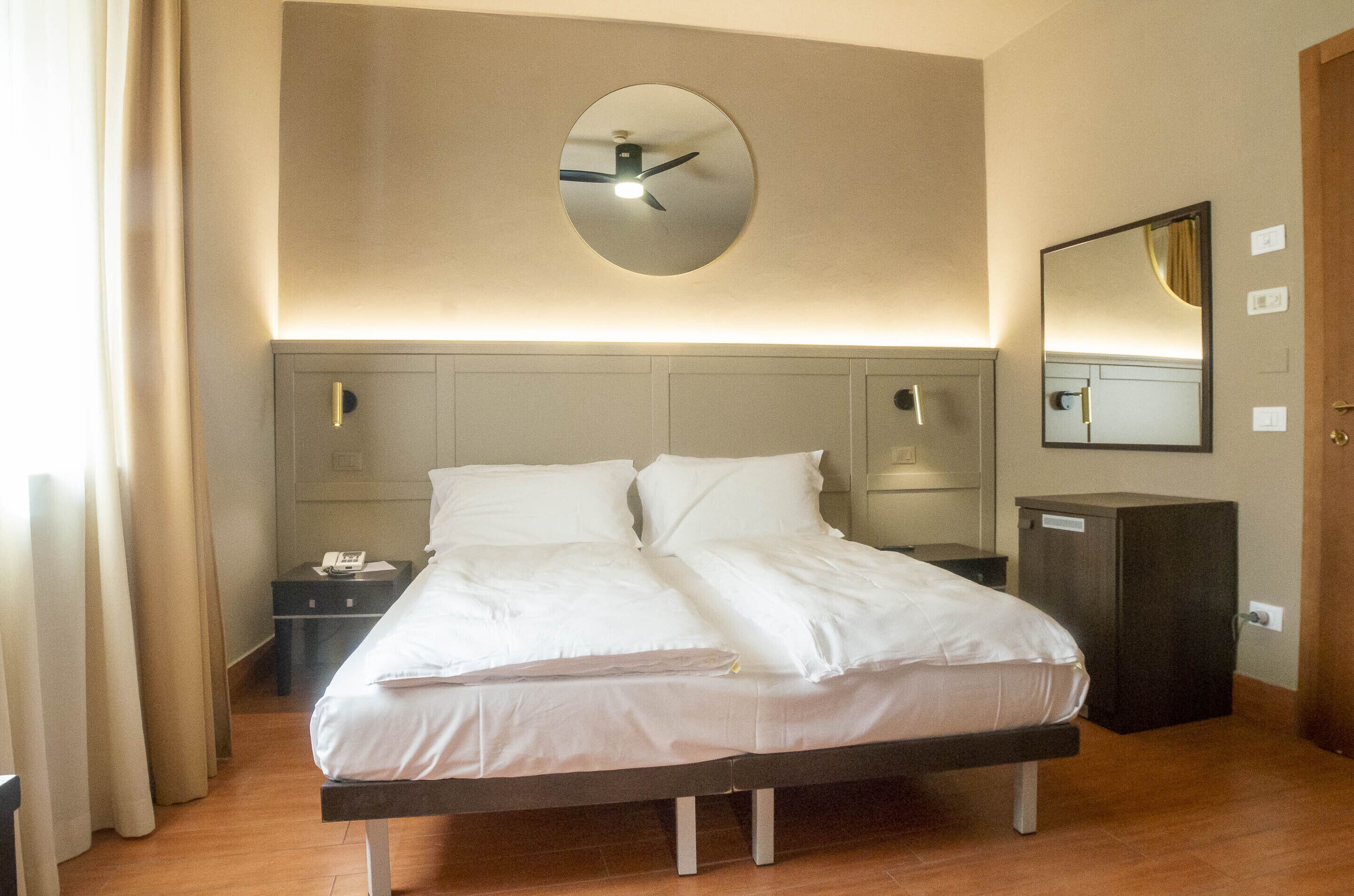 A hotel room with a bed, bedside tables, a mirror, and a clock on the wall.