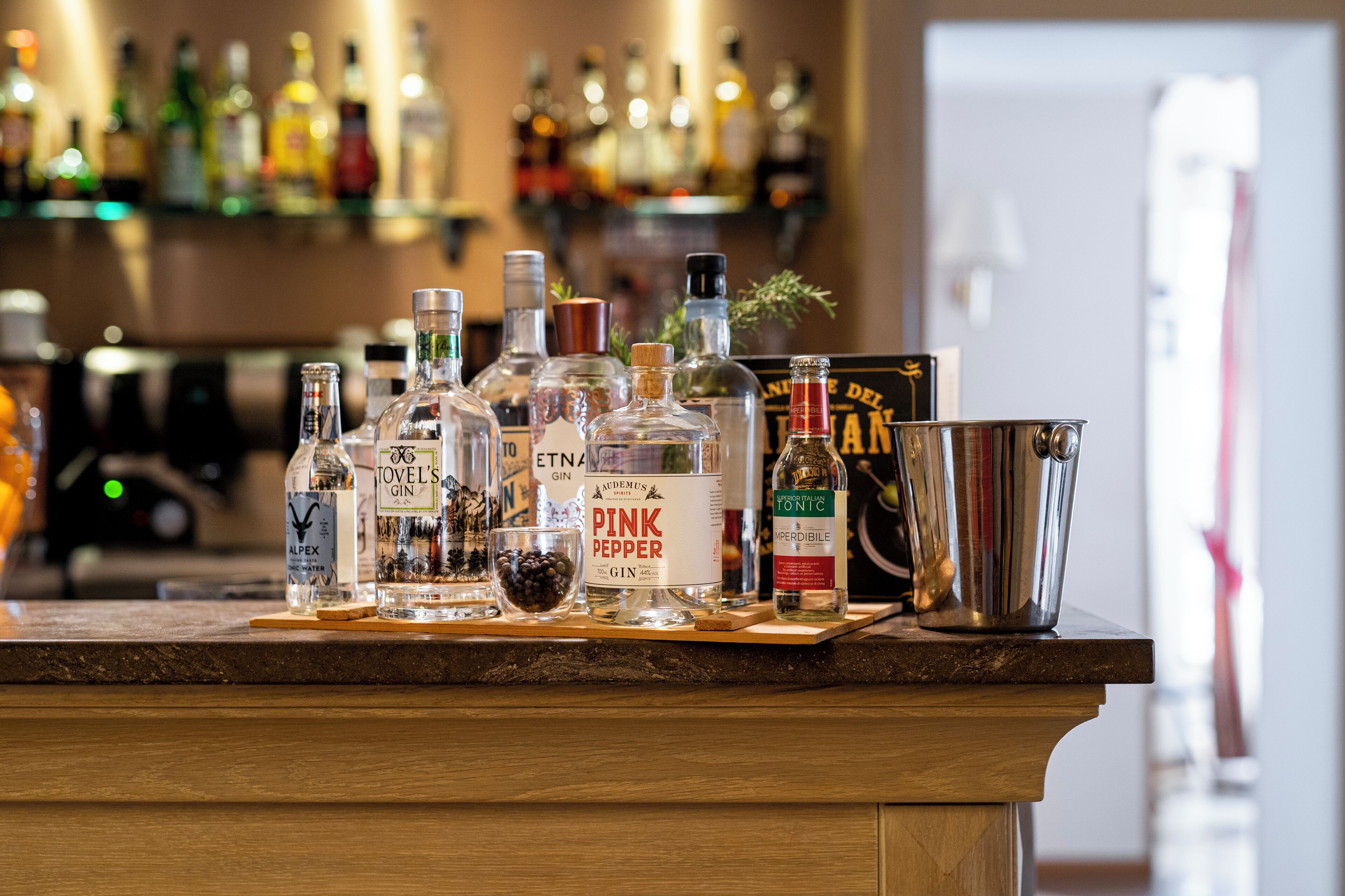 Et barområde med forskellige flasker alkohol, heriblandt gin og whiskey, samt en metalkværn på en træbordplade.