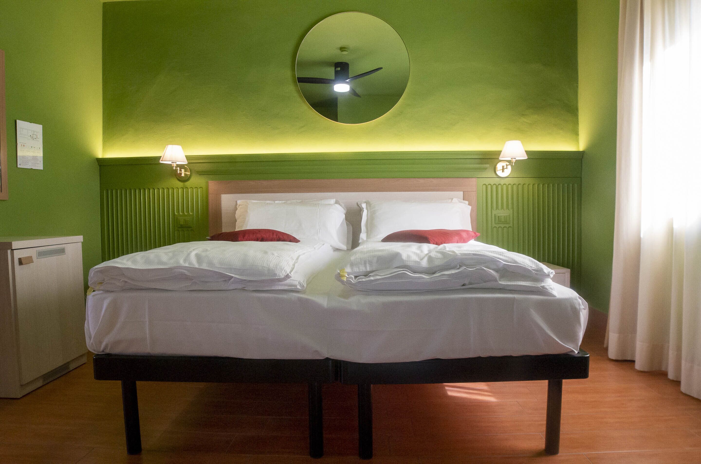 A double bed with white linens and red pillows, a round mirror above, two wall-mounted lamps, and a radiator below.