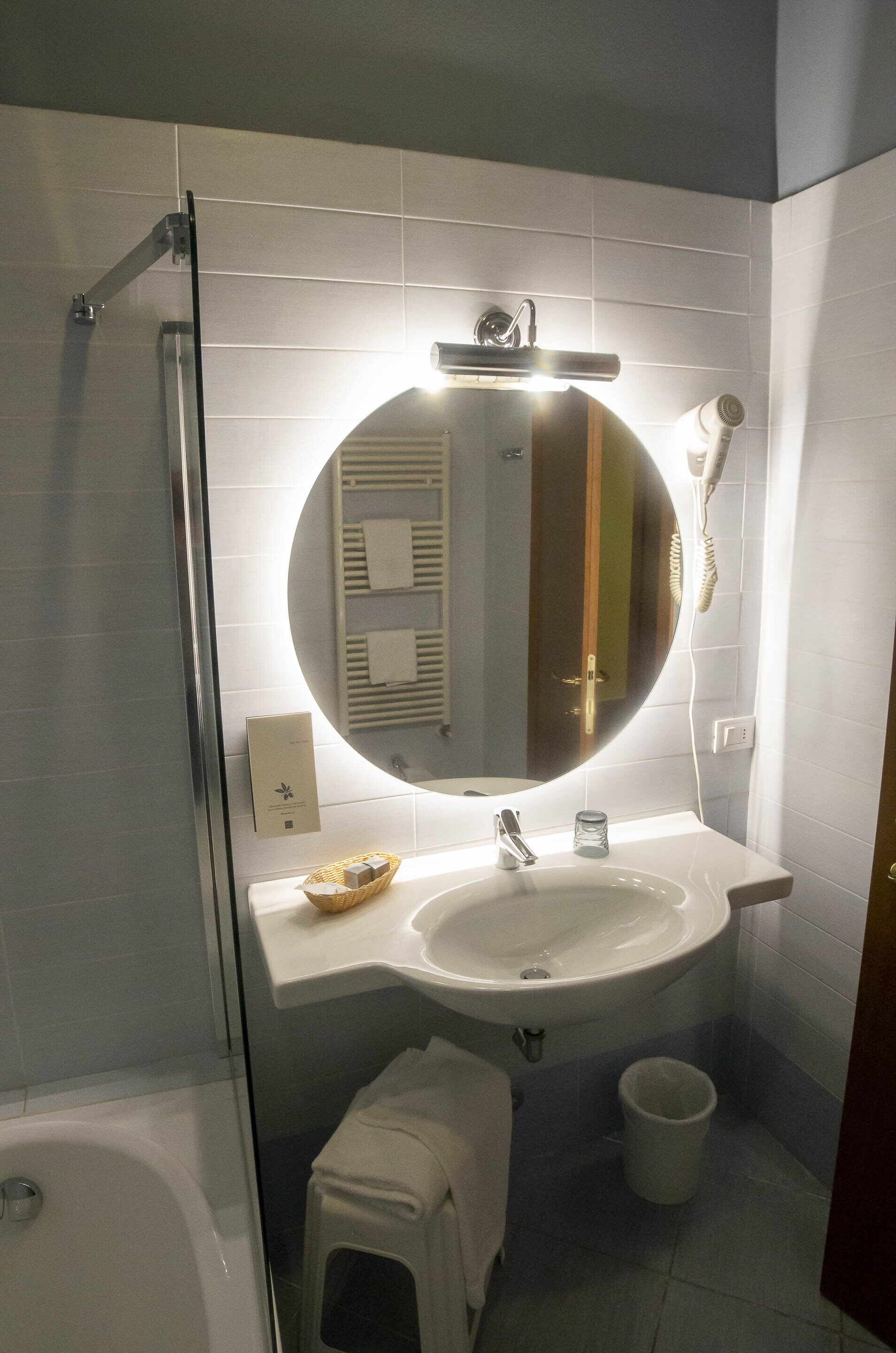 Een badkamer met een ronde spiegel, een wastafel, een losse haardroger en een klein prullenbakje.