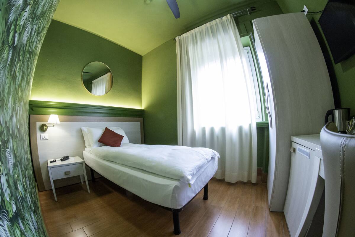 A hotel room with a bed, a nightstand, a lamp, a mirror, a window with curtains, and a small kitchenette.