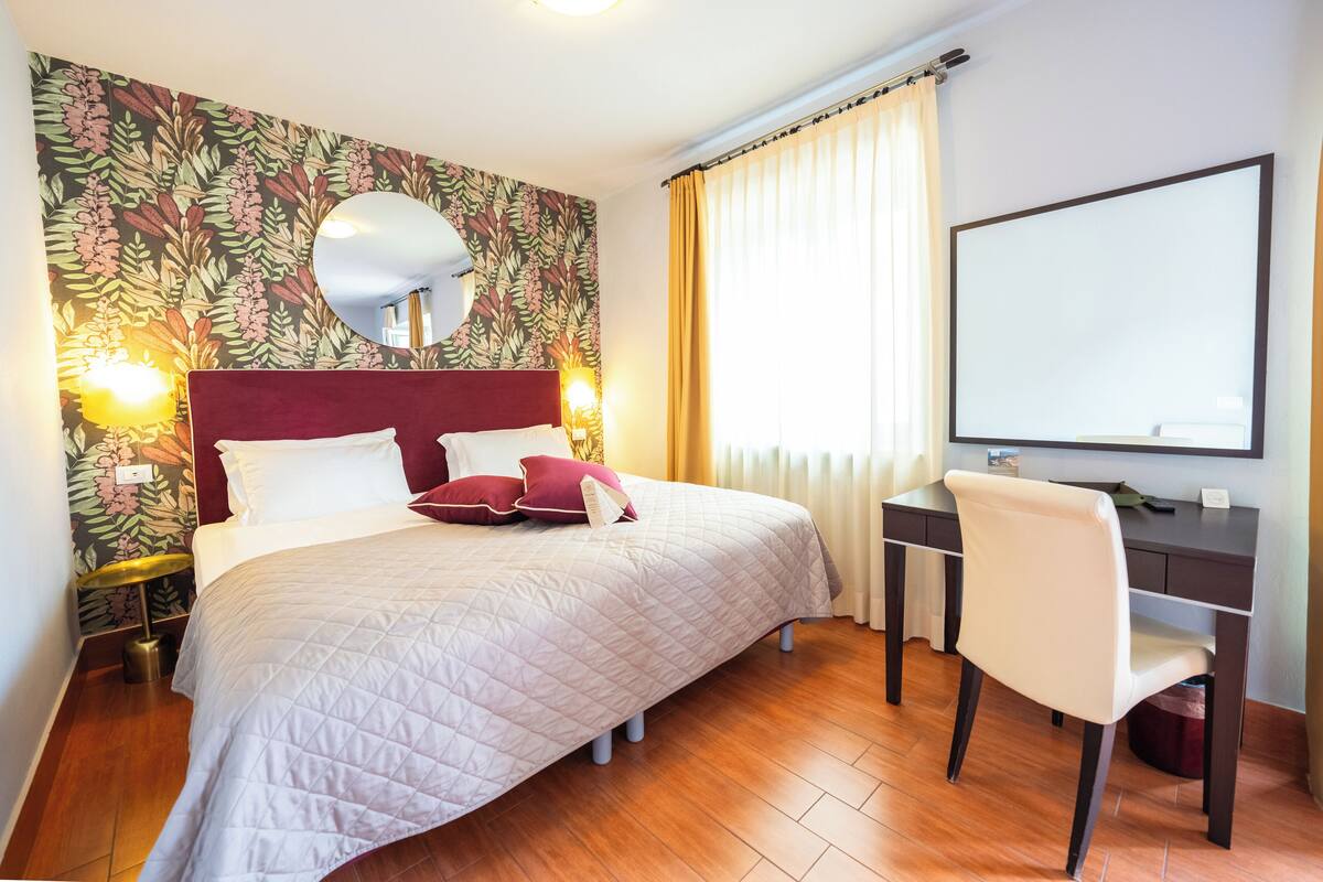 Habitación de hotel con cama, escritorio, silla, espejo y un amplio ventanal con cortinas.