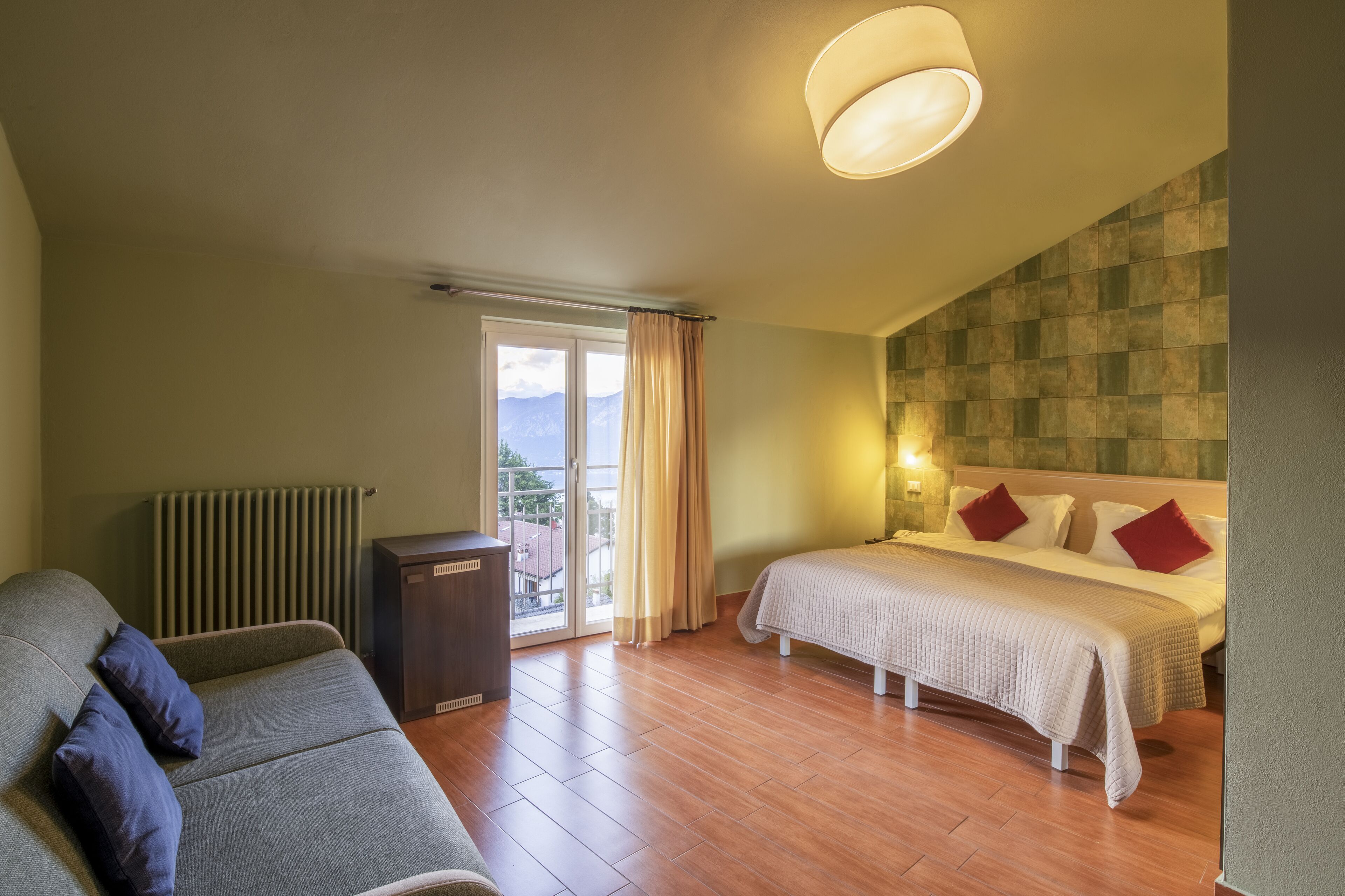 A hotel room with a bed, a sofa, a radiator, and a view of the outdoors.