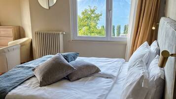 Una cama bien hecha con sábanas y almohadas blancas, un espejo redondo en la pared, una ventana con vistas a montañas y un radiador.