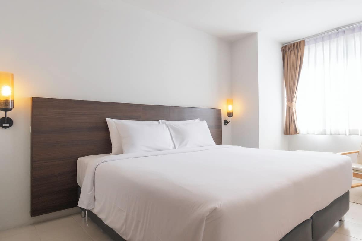 A hotel room with a large bed, two wall-mounted lights, a wooden headboard, a chair, and a window with curtains.