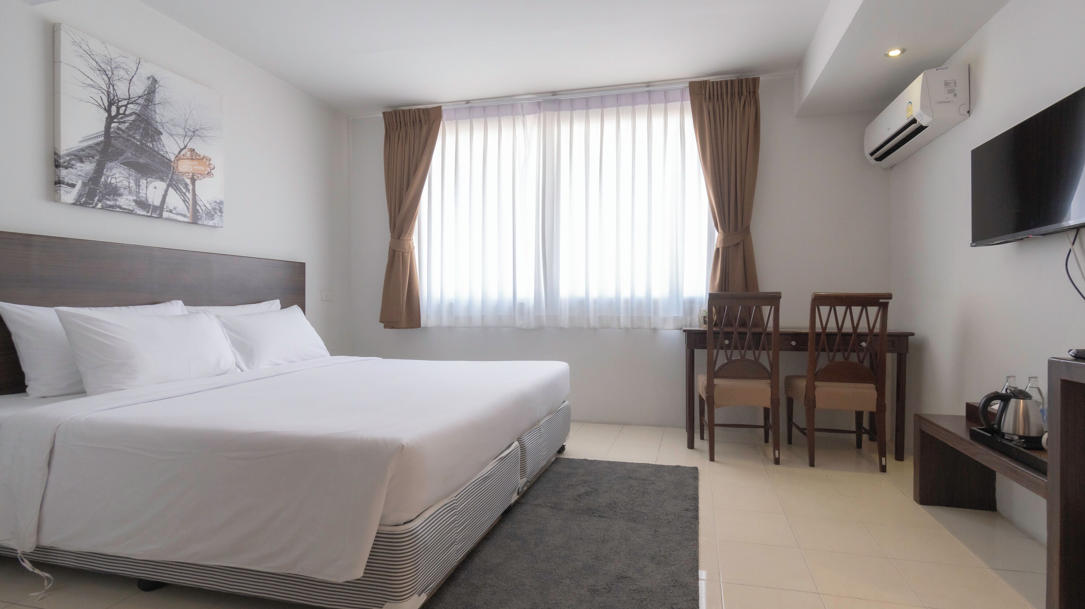 A hotel room with a bed, a desk with two chairs, a television, and an air conditioning unit.
