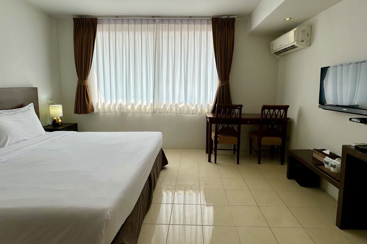 A hotel room with a bed, a desk with chairs, a television, and an air conditioning unit.