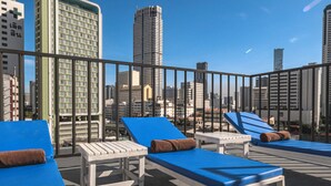 Un salón en la azotea con sillones azules y mesas blancas, con vistas a un paisaje urbano con rascacielos.