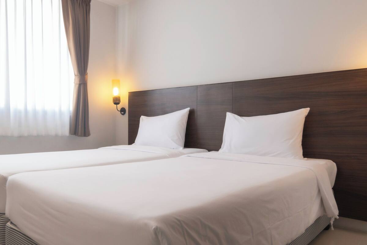 A hotel room with two beds, a wooden headboard, a wall-mounted light, and a window with curtains.