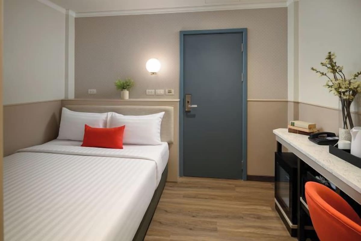 A hotel room with a bed, a desk, a chair, a door, and a wall-mounted light.