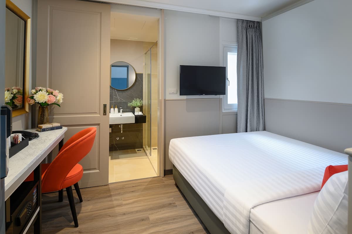 A hotel room with a bed, a desk with a chair, a TV, and a bathroom visible through an open door.