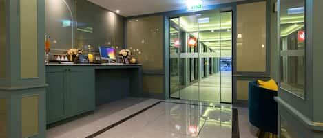 A hotel lobby with a reception desk, glass doors leading to a hallway, and a blue chair.