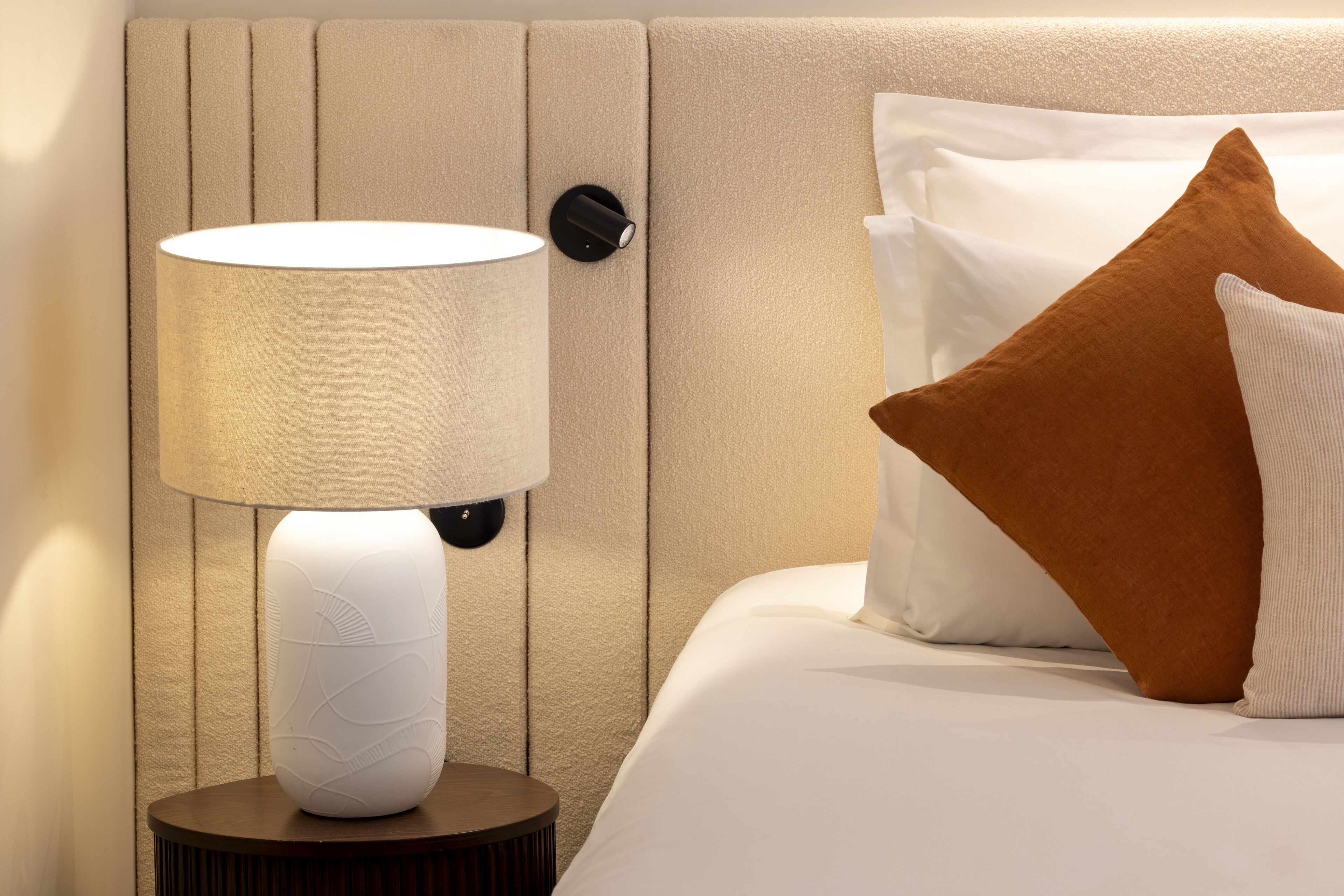 A neatly made bed with white and brown pillows, a round bedside table with a white lamp, and a textured wall panel.