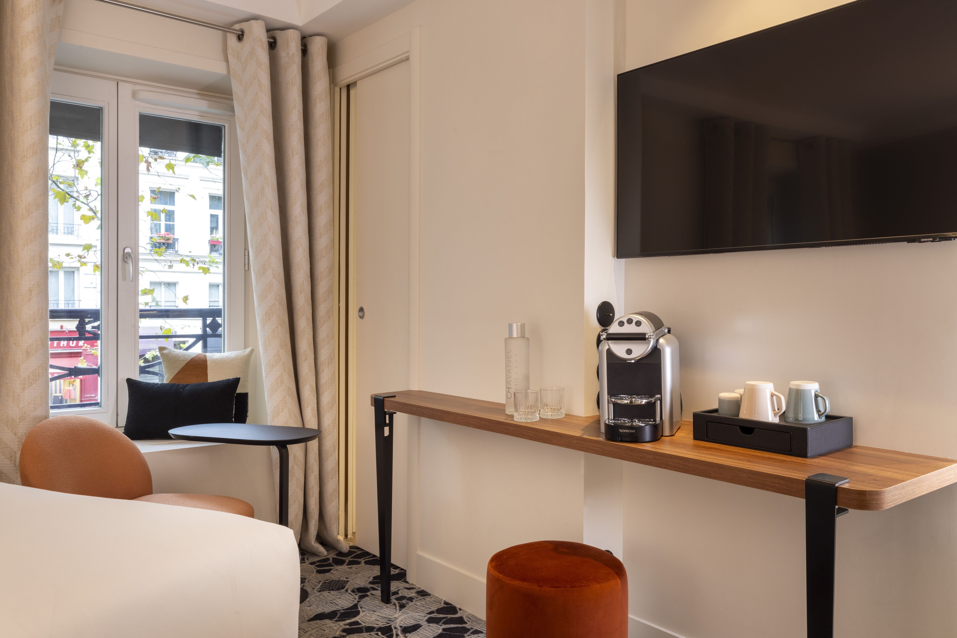 Quarto de hotel com cama, uma mesinha com cafeteira, televisão na parede e janela com cortinas.