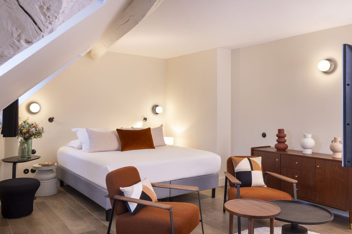 A modern bedroom with a bed, two chairs, a small table, a dresser, and wall-mounted lights.